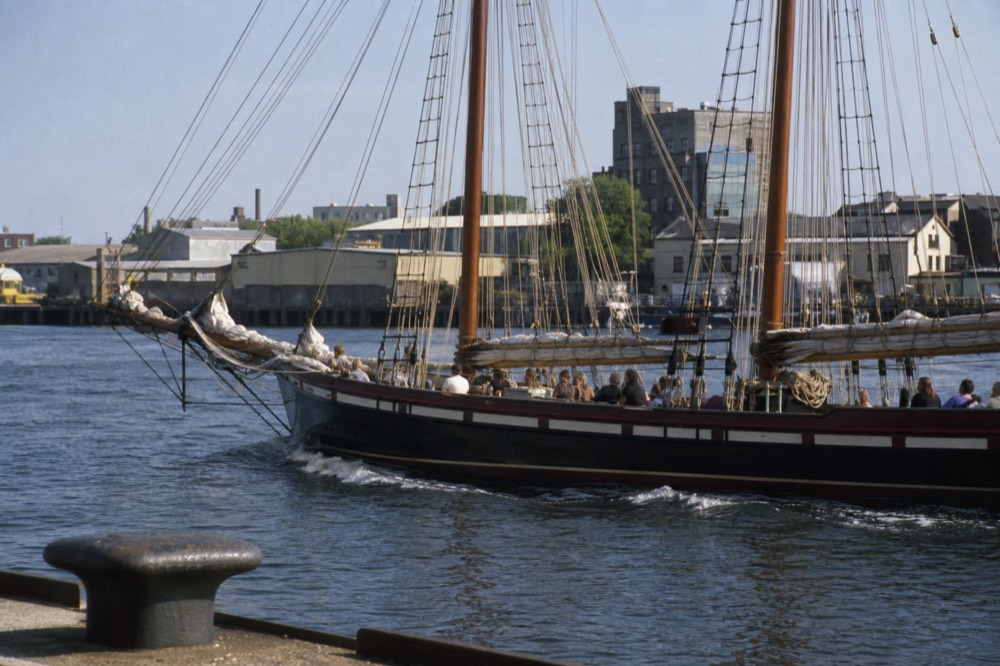 Tall sailing ship with passengers seated along a high dry deck on harbor water, no text, no logos, no people.