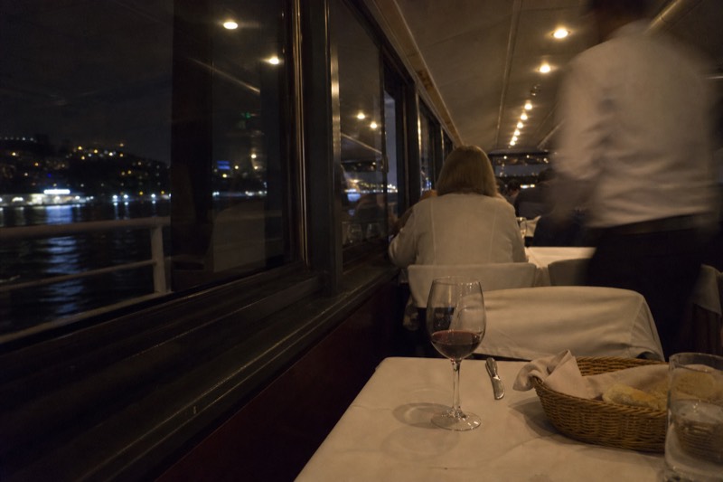 Dinner tables set on a covered deck at night on a Charleston harbor dinner cruise, city lights reflecting on the water