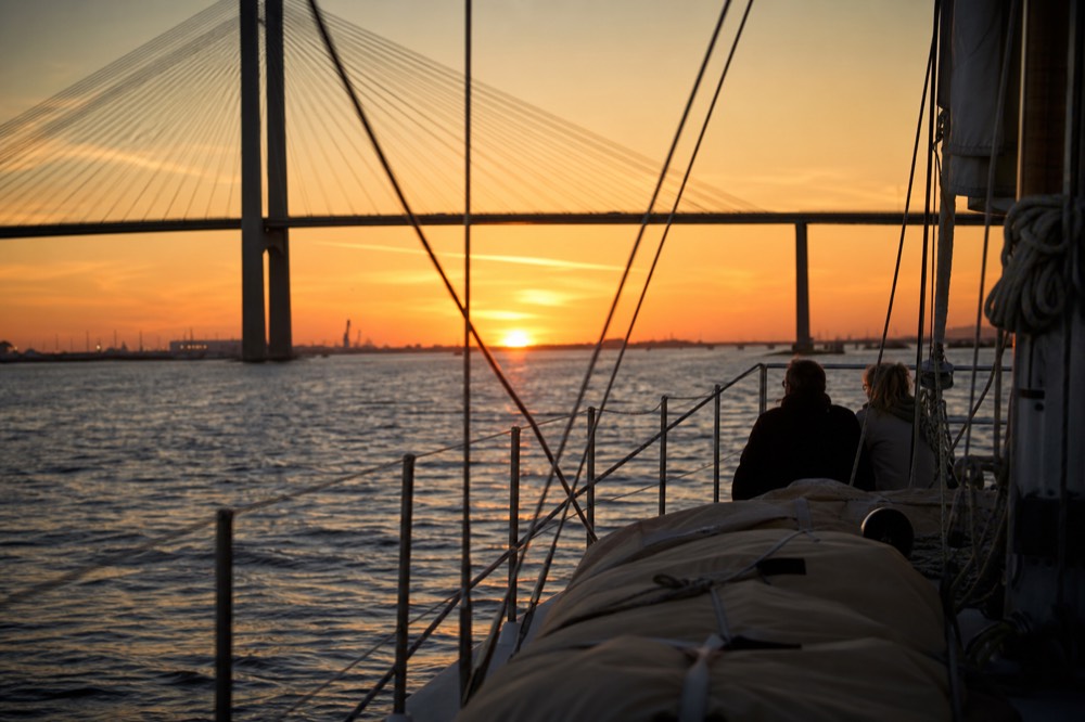 The sun setting behind the Ravenel Bridge as a sailing cruise crosses the harbor, no text, no logos, no people.