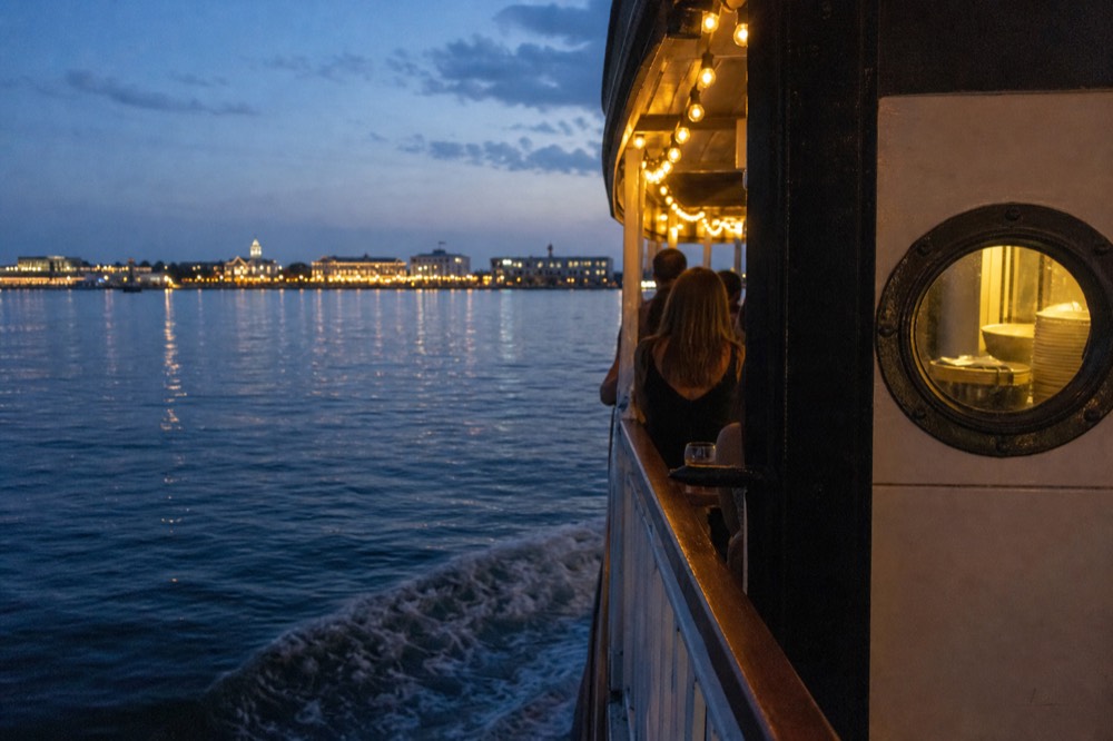 A dinner cruise boat lit for the evening departing past the Charleston waterfront, no text, no logos, no people
