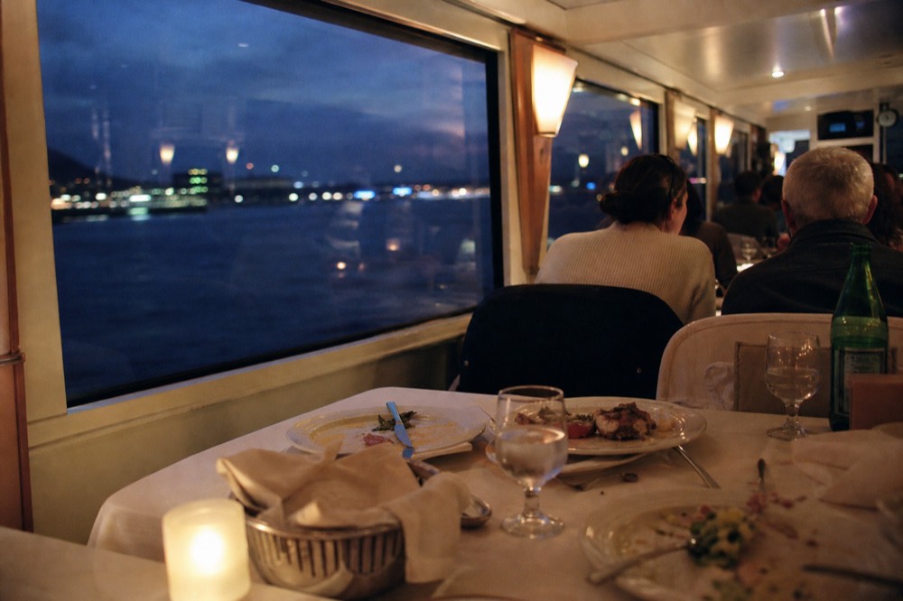 Guests seated at a set dinner table aboard a cruise boat with harbor views through large windows, no text, no logos, no people