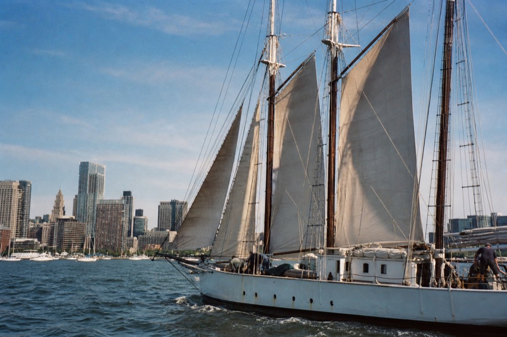 A three-masted schooner under sail on Charleston harbor with the city skyline behind, no text, no logos, no people