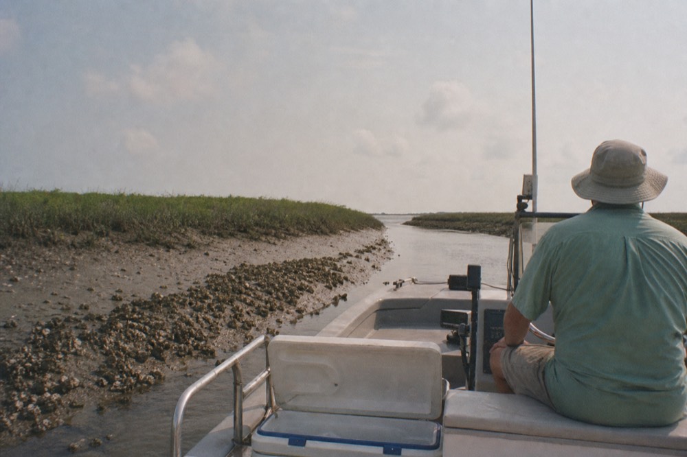 Small tour boat drifting through a narrow salt marsh creek at low tide, no text, no logos, no people
