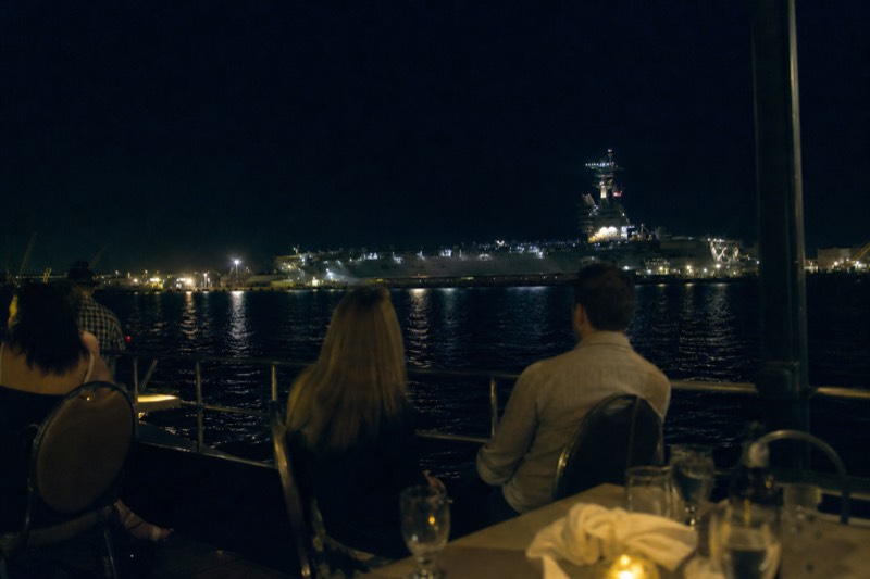 Charleston harbor dinner cruise boat with live music, windows glowing as it passes the lit aircraft carrier at night