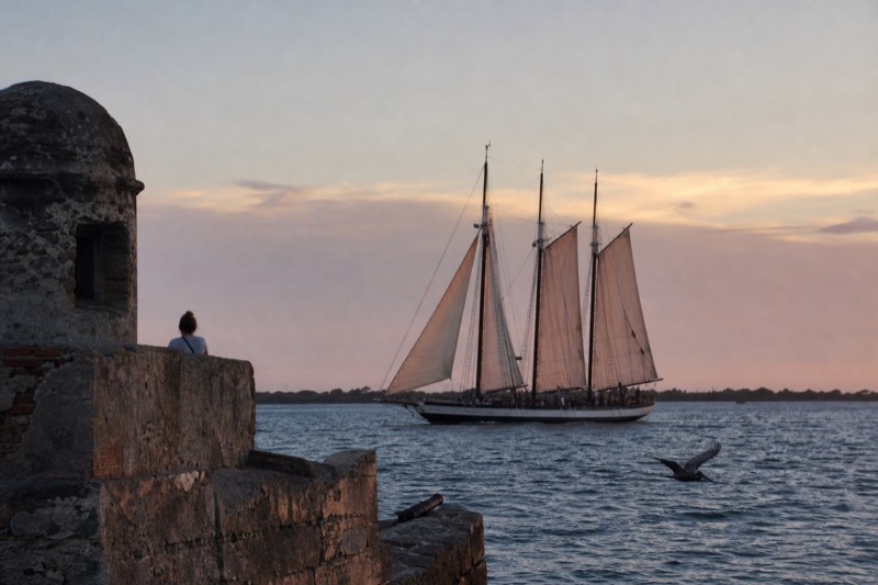 Three-masted schooner under full sail gliding past a Charleston harbor fort at dusk